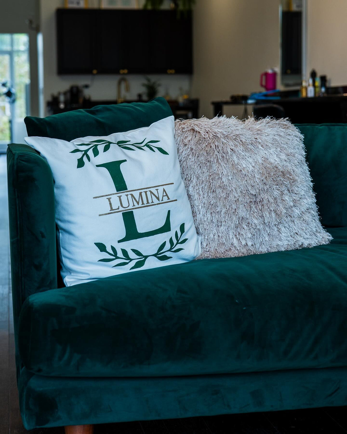 Green velvet sofa with a Lumina monogram pillow in the salon lounge at Lumina Extensions & Beauty Bar, Cary NC