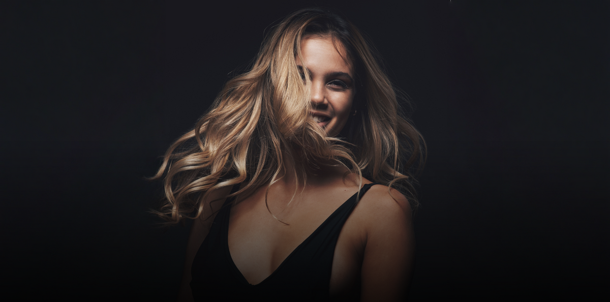 Woman with long wavy blonde hair tossing her hair in dark studio light at Lumina Extensions & Beauty Bar, Cary NC